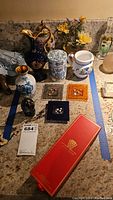 Photo showing assorted decorative items including Limoges vase, ceramic jar and pitcher, yellow flower figurines, small dark vase, three square art glass plates with button accents and a long red box with a white butterfly hanging ornament.