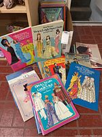 Photo showing a stack of vintage paper doll books with illustrated covers including Opera Stars of the Golden Age, and Review Madame Alexander Doll magazines spread out on the floor.