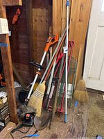 Photo showing Black & Decker weed wacker leaning against wooden wall along with brooms, shovels, other garden tools and hardware items on wooden floor