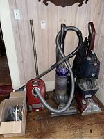 Lot of three upright vacuum cleaners together with Dyson in center, Hoover and Miele on sides, plus box with vacuum parts.