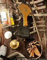 Top-down view showing metal log grates, a wooden bellows, oil lamp with brass lid, black ash buckets filled with kindling wood, fire starters, and safety matches on a brick floor.