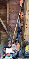 Photo showing multiple Black & Decker garden power tools including two hedge trimmers, a leaf blower, and accompanying accessories leaning against a wooden wall.
