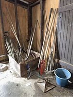 Photo showing several garden and yard tools grouped together in a corner including rakes, shovels, diggers, and a blue bucket, all with wooden handles and dirt on them.