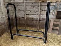 Metal log rack frame standing upright on straw-covered floor in a barn or shed. Black tubular metal with two vertical frames and horizontal support bars.