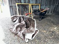Two yellow adjustable metal saw horses leaning against barn wall near black wheelbarrow, brown tarp on gravel ground, and driveway markers inside wheelbarrow