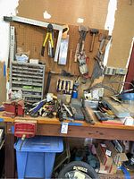 Workbench with attached vice, hand tools on surface, pegboard overhead with saws and hammers, hardware organizer