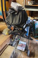 Full view of Weber grill with black cover, propane tank beneath, set on wheeled cart indoors.