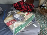 Full assortment on bed: buffalo plaid blanket on top, packaged striped and cream wool blankets beneath, Samsonite roll on top