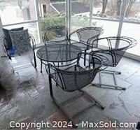 Five black wrought iron chairs with mesh seats and backs surrounding a glass table in an enclosed porch area.