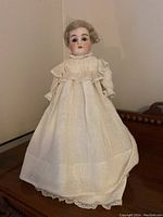 Full frontal view of doll standing on wooden surface in white lace dress.