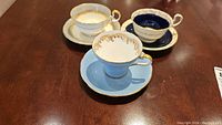 Three Aynsley teacups and saucers on wooden table showing different styles and colors.