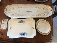 Four bone china serving pieces on wood table, includes one long gold gilt tray and smaller blue and white and gold trimmed dishes