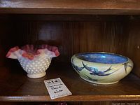 Two bowls side by side on wooden shelf: ceramic Grimwade Swallows bowl with bird pattern and crazing on right, white hobnail Fenton bowl with pink ruffled rim on left.