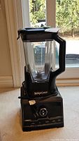 Front view of Ninja Chef blender showing blending container, base, and power cord. Blender is black with clear container and measurement markings.