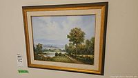 Framed vintage landscape painting showing countryside road, trees, house, and water body.