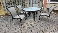 Photo of round outdoor table with frosted glass top and four matching chairs with metal frames and mesh fabric seats and backs, placed on a paved patio.