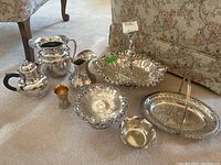 Full view of all silverplate serving ware including two jugs, small bowls, footed dish, basket dishes, and a shot cup arranged on carpet near furniture