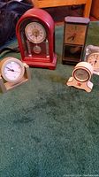 Group image showing five clocks on a green surface, various sizes and designs including red wooden framed pendulum clock and smaller beige clock.