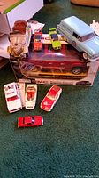 Photo showing various diecast and toy cars arranged on a surface next to their original boxes; displays a variety of brands and vehicle types, colors red, white, green, yellow, and blue