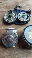Three Orvis fly fishing reels and a zippered case laid on wood surface.
