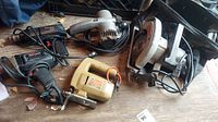 Top-down view of laid out power tools including circular saw, jig saw, two electric drills, and belt sander on wooden surface