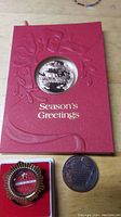 Franklin Mint Season's Greetings coin in red booklet with two bronze-tone commemorative medals on wooden table.