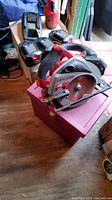 Photo showing the 18V Skilsaw circular saw with attached blade, three batteries, and battery charger on and near a filing box case