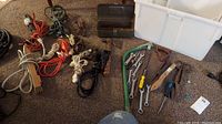 Photo of mixed extension cords in various colors, a small metal toolbox, hand tools including wrenches, saw, cutters, brush, and the white plastic tote with lid on carpeted floor.