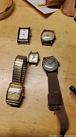 Five vintage wristwatches displayed on wooden surface: Tourneau with gold-tone metal band; Bijoux-Terner small watch without band; Helbros wind-up watch with worn dial; Stellaris watch without band or pins; Hamilton watch with gold-tone band and wear.