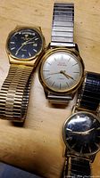 Four wristwatches placed on a wooden surface, showing the Jules Jürgensen quartz watch with gold-tone face and band, Elgin Sportsman with silver face and metal band, and two other watches including one with a dark face and gold bezel.