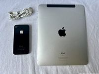 Overview of the Apple iPhone 4, Apple iPad 1st Gen, and USB charging cable laid flat on a white cloth background, showing backs of devices.
