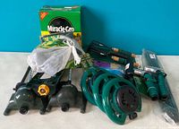 Photo showing assorted gardening items including a green Miracle-Gro box, Orbit sprinkler heads, green coiled hose, and hand gardening tools.