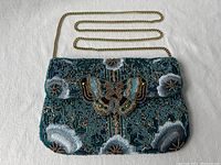 Front view of blue beaded handbag with butterfly and floral design and gold chain strap laid flat on white fabric.