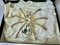 Large rhinestone spider brooch lying against white fabric inside a box, showing full front view.