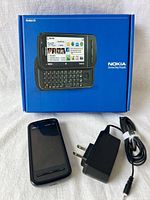 Nokia C6 phone in front of its original blue retail box with slide-out QWERTY keyboard shown on the box. Black charger with plug and cable is laid beside the phone.