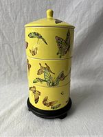 Front view of the assembled famille jaune porcelain carrier with three stacked round boxes and fitted lid on black wooden stand. Yellow background with multi-colored butterflies.