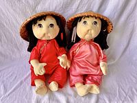 Pair of two vintage soft sculpture dolls styled as Asian rice paddy babies, one in red outfit, one in pink outfit, both wearing woven straw hats, seated side by side on white fabric background.