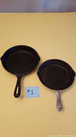 Two cast iron frying pans side by side, one black and one silver-gray, shown with measuring tape and reference number