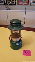 Full view of green Coleman Lantern Model 335 with carry handle and red valve knob on yellow table.