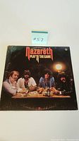 Front of the vinyl album cover showing the band Nazareth seated at a table with drinks and cigarettes, album title 'Play 'n' The Game' in orange on top.