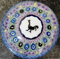 Top view of vintage Baccarat glass paperweight showing black rooster silhouette in center with surrounding colorful millefiori floral design under clear glass dome.
