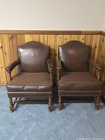 Pair of H-Krug arm chairs next to wooden wainscoting wall, showing front view with brown leather backs and fabric cushions.