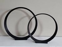 Two black metal ring sculptures shown side by side on a white surface against a light background, circular shape with flat base.