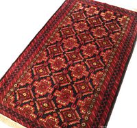 Angled view of the full Baluch rug showing intricate geometric pattern in cherry red and black with beige highlights and fringes on the edges.