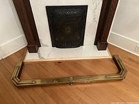 Full view of brass fireplace surround positioned on wooden floor near fireplace opening, showing overall condition and shape.