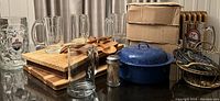 Wide angle showing glass mugs, wooden utensils and cutting boards, blue enamel pot, metal trivet, and cardboard boxes.