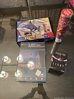 Lot of assorted kids games and toys arranged on glass surface including remote control flying shark, LED Empire State Building 3D puzzle, plasma orb light, and flying discs canister.