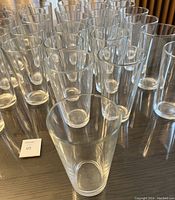 Photo shows 30 clear cylindrical beer glasses arranged on a dark surface. Glasses are transparent and uniform in size and shape.
