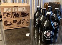 Photo showing BrewShop How To Brew beer making kit box with illustrated instructions on front and five Parallel 49 branded brown glass growler jugs with white caps with handles.