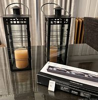 Two black metal lanterns on a table, each containing a beige pillar candle. One lantern has cracked glass. A boxed Candlescape set is on the table beside the lanterns.